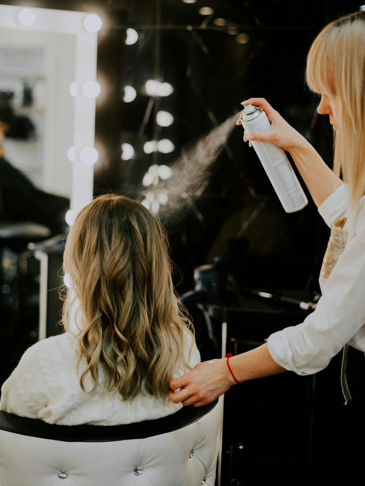 Uma cabeleireira aplica um produto de acabamento em spray sobre o cabelo de uma cliente após um balayage, a secagem e a modelagem em ondas