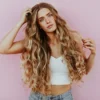 Foto de estúdio de uma mulher com o cabelo comprido, ondulado e beijado pelo sol com balayage, vestida com calças de ganga azul-claro e um top curto branco de malha