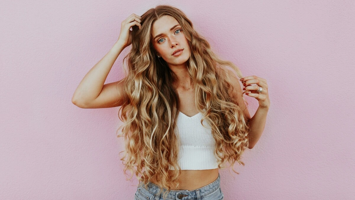 Foto de estúdio de uma mulher com o cabelo comprido, ondulado e beijado pelo sol com balayage, vestida com calças de ganga azul-claro e um top curto branco de malha