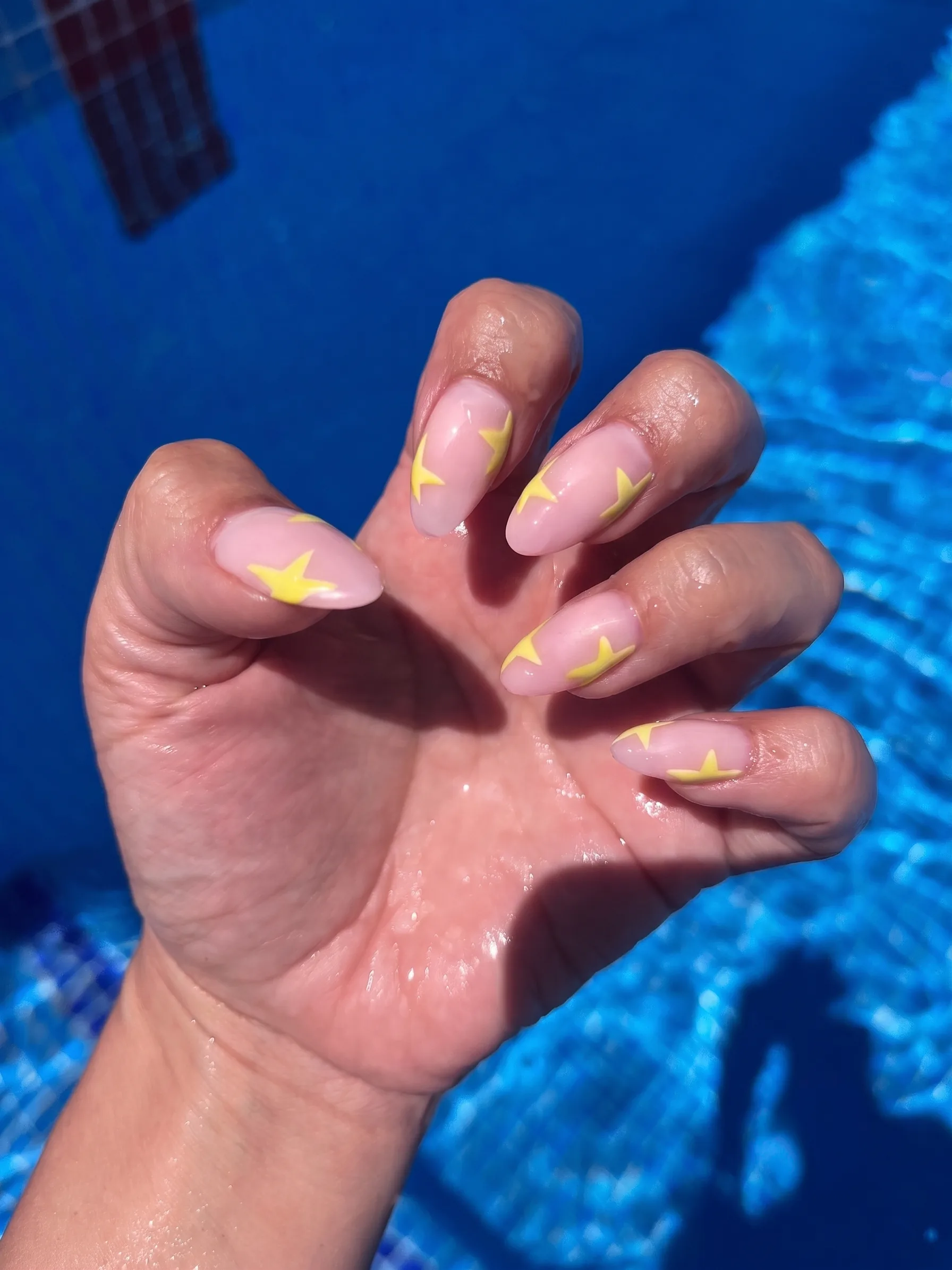 Unhas em formato amêndoa em nude transparente com estrelas em amarelo manteiga, fotografadas à beira da piscina.