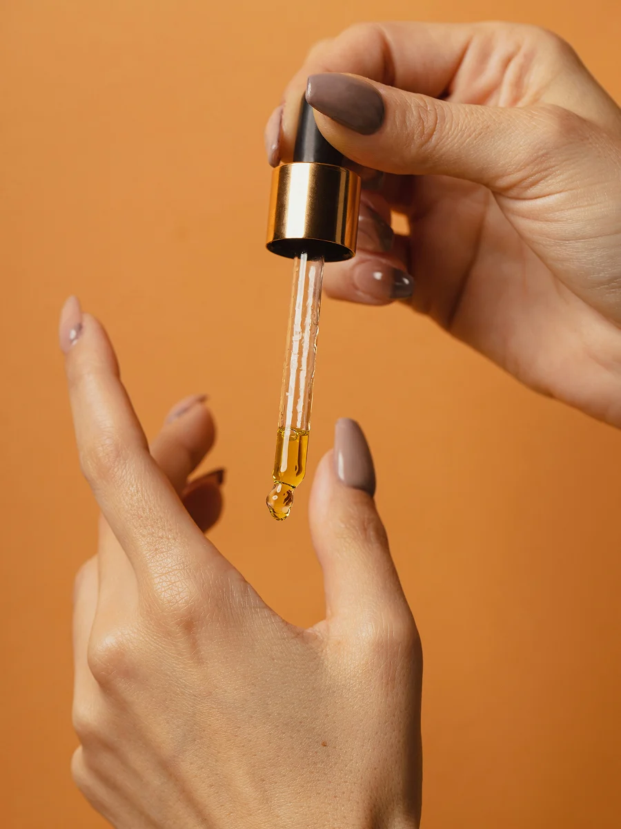 Grande plano de duas mãos com manicure, uma delas a segurar um conta-gotas de vidro com óleo de cutículas a escorrer