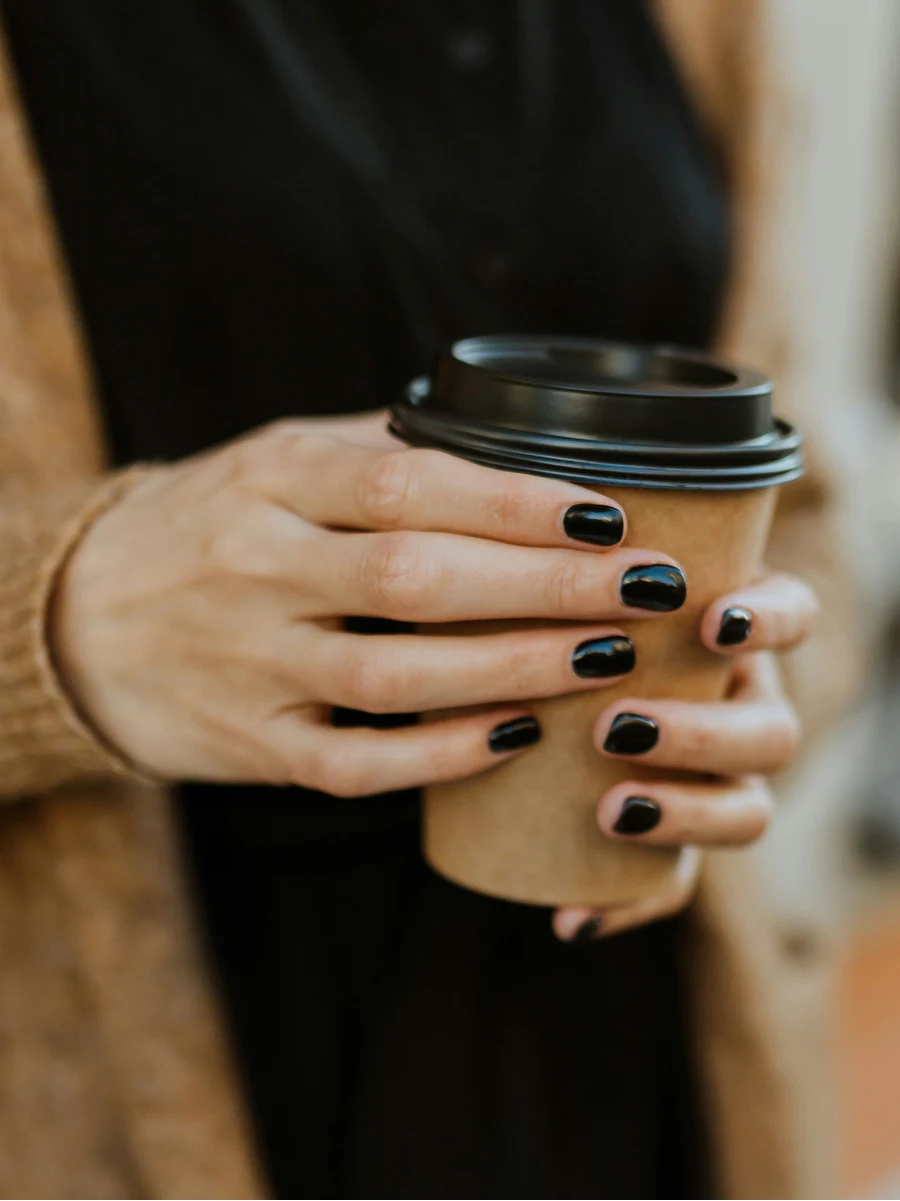 Grande plano de uma mulher com uma manicure em gel preto brilhante, a segurar um copo de café para levar