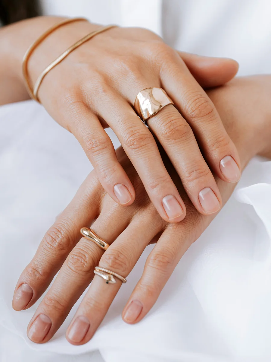 Grande plano de duas mãos pousadas sobre um lençol branco, com unhas naturais e saudáveis e adornadas com anéis e pulseiras em rosa-dourado