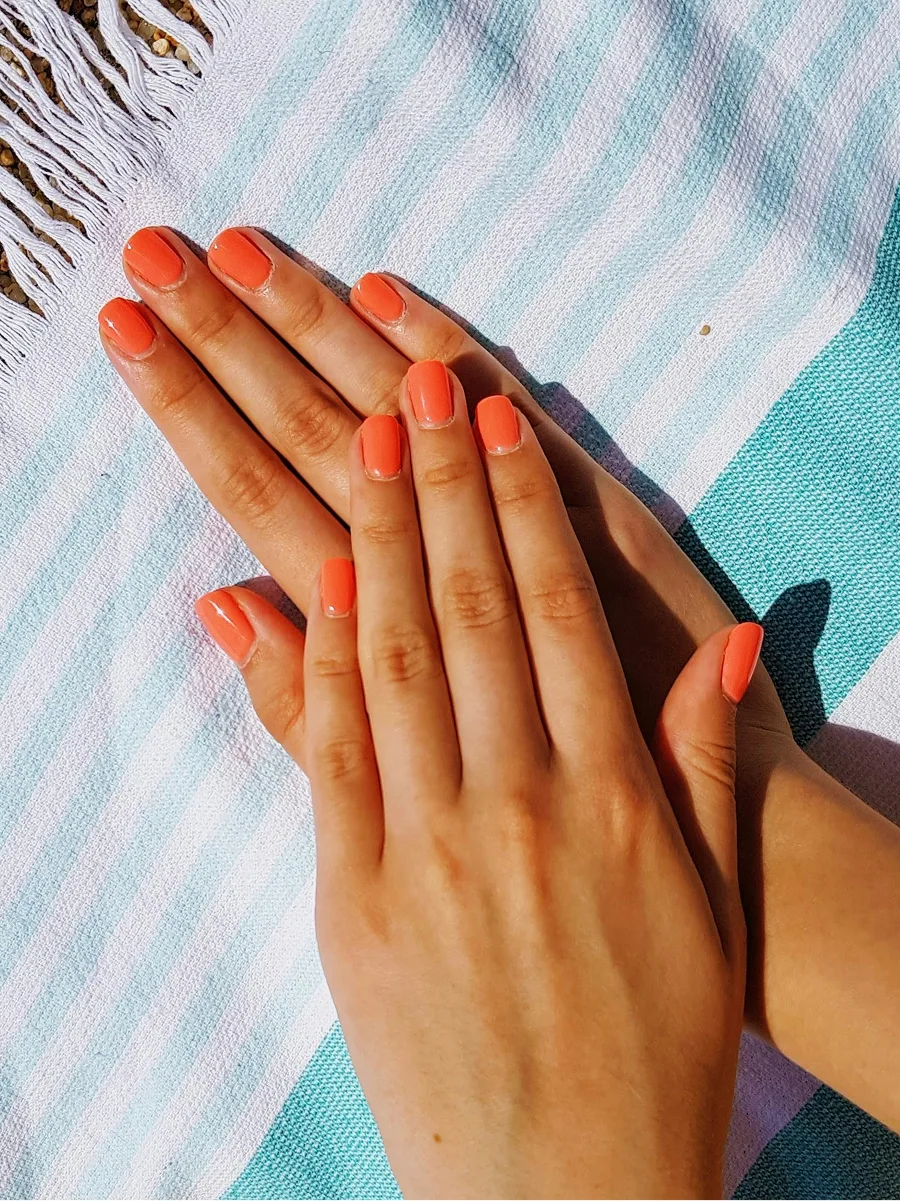 Vista de cima de duas mãos com uma manicure em gel laranja brilhante, pousadas sobre uma toalha de praia às riscas azuis e brancas