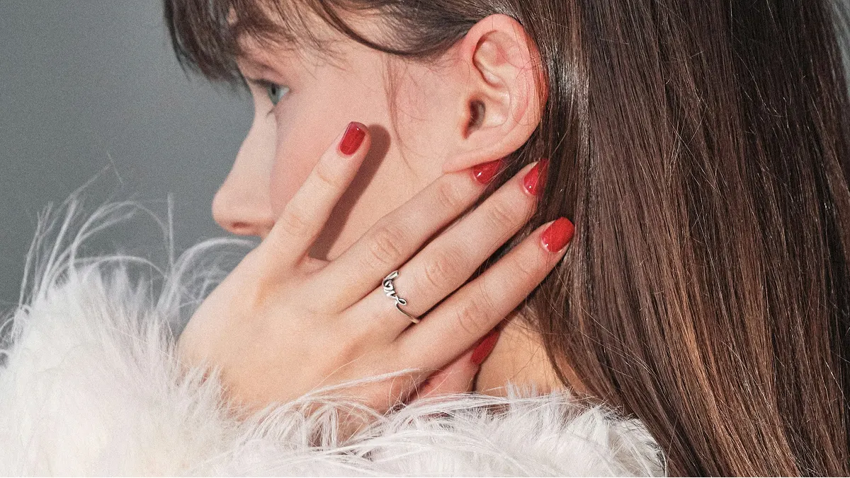 Vista lateral em grande plano da cabeça de uma mulher morena, com a mão a tocar no rosto e a exibir uma manicure em gel vermelho brilhante