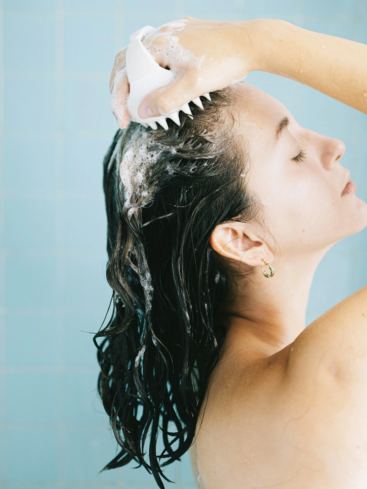 Vista lateral de uma mulher com o cabelo molhado e ensaboado a massajar o couro cabeludo com uma escova de silicone