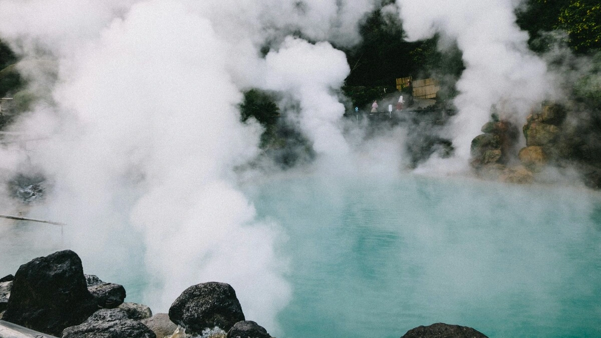 Vista ampla de termas no Japão com nuvens de vapor a subir de um tanque rodeado de rochas e vegetação