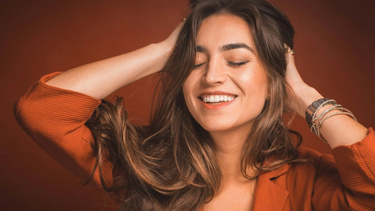 Grande plano de uma mulher a sorrir de olhos fechados enquanto passa as mãos pelo seu cabelo castanho, comprido e macio