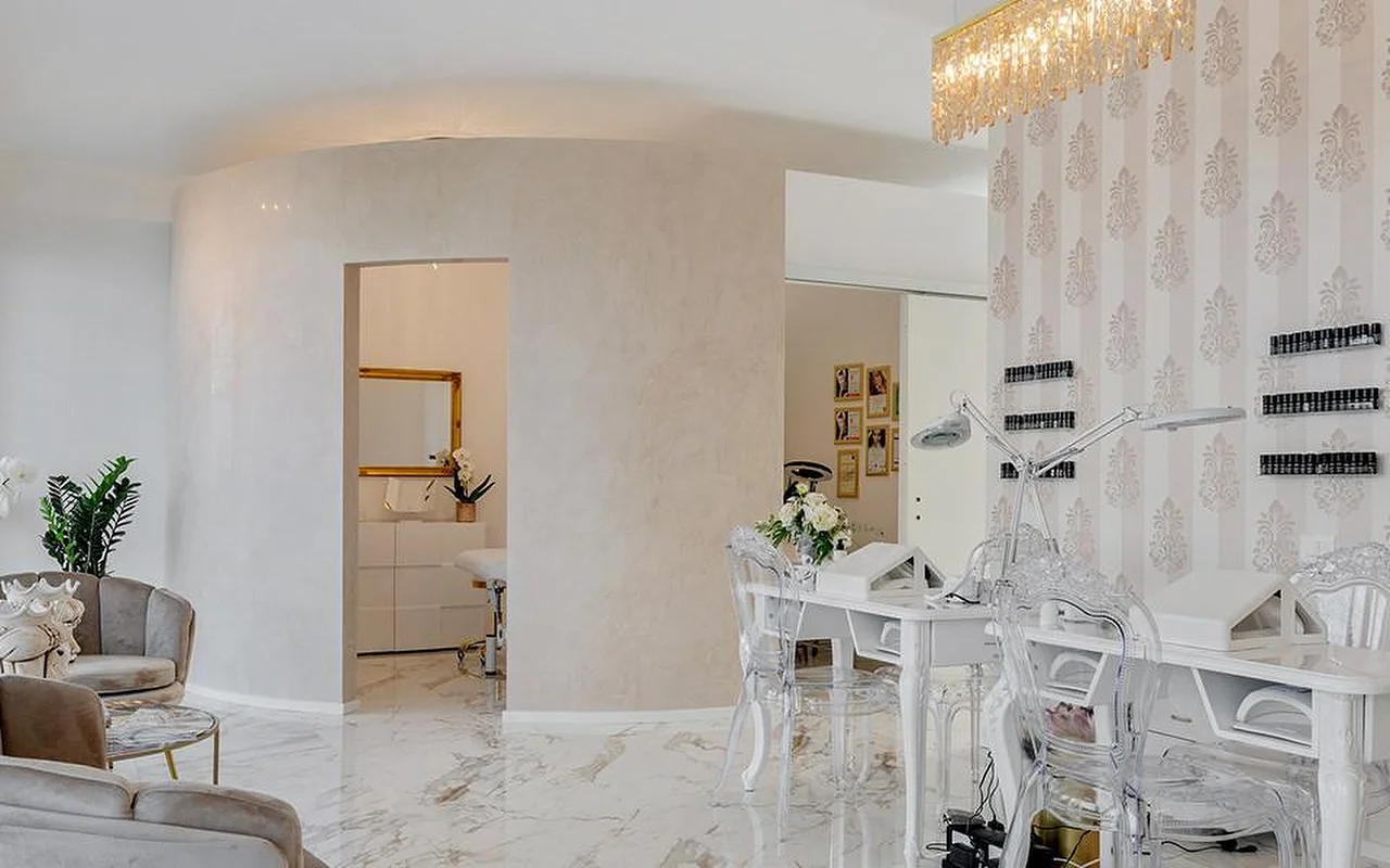 O interior elegante do MC Beauty Center com pavimento com efeito mármore, paredes curvas brancas e um candeeiro de teto em cristal dourado. Uma estação de unhas com cadeiras transparentes fica à direita, enquanto poltronas de veludo e uma porta com moldura dourada dão para uma sala de tratamento ao fundo.