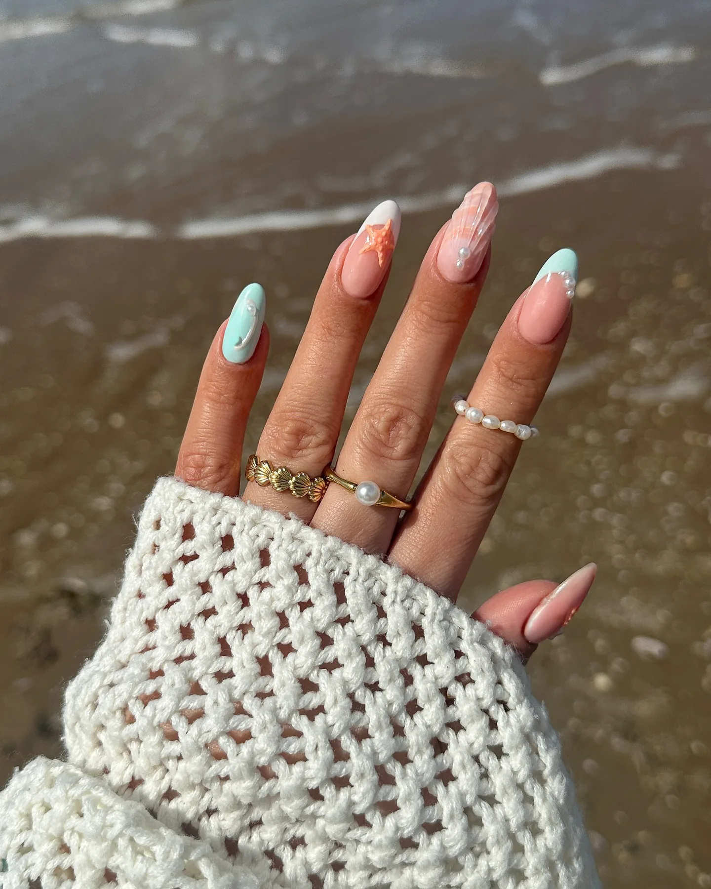 Unhas em formato amêndoa com nail art de sereia em tons pastel – uma estrela-do-mar, apontamentos de madrepérola e um brilho iridescente – sobre areia molhada.