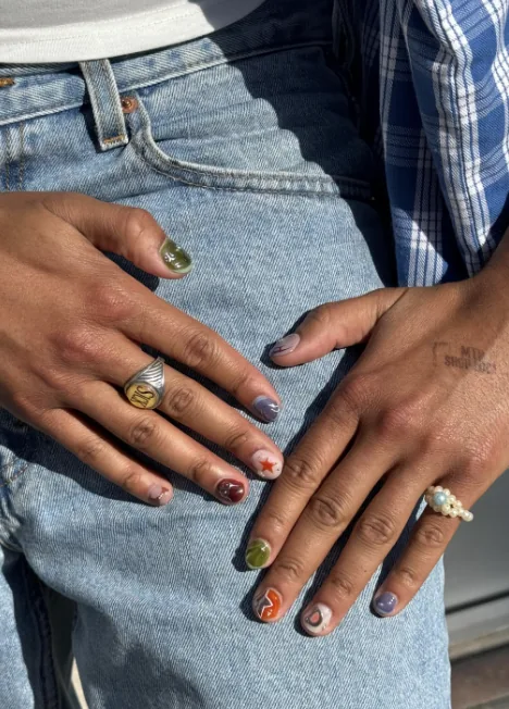 Manicure a descombinar em unhas curtas, cada unha com uma cor e um desenho diferentes, com uma estrela vermelha e um floreado laranja, pousada sobre umas calças de ganga.