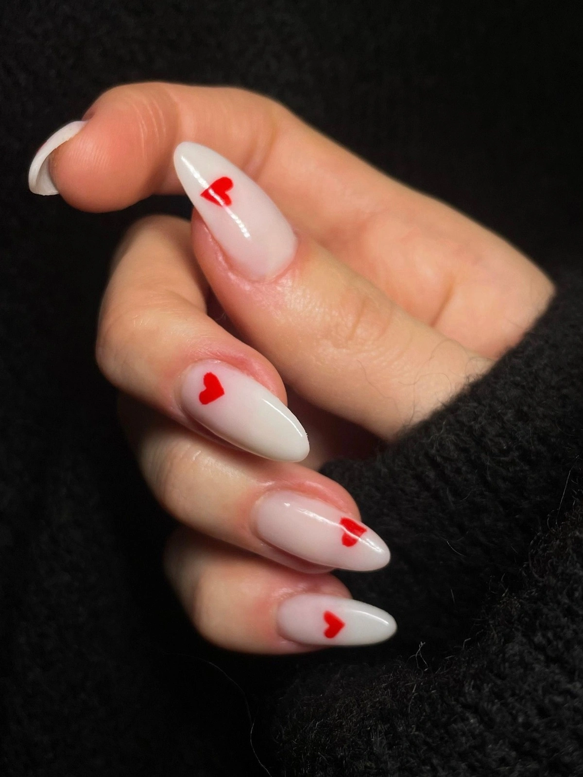 Grande plano da mão de uma mulher com unhas de acrílico brancas em formato amêndoa, decoradas com pequenos corações vermelhos