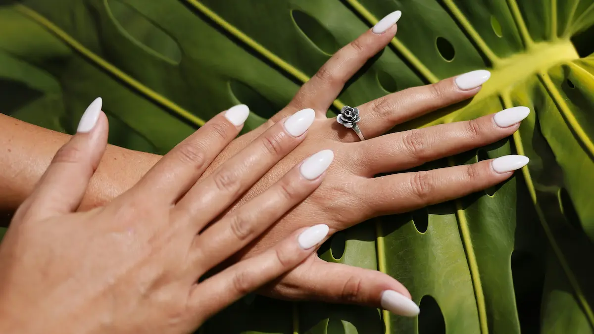 Fotografia de estúdio de unhas de acrílico acabadas de fazer, em formato amêndoa e tom nude, sobre um fundo de folha de palmeira
