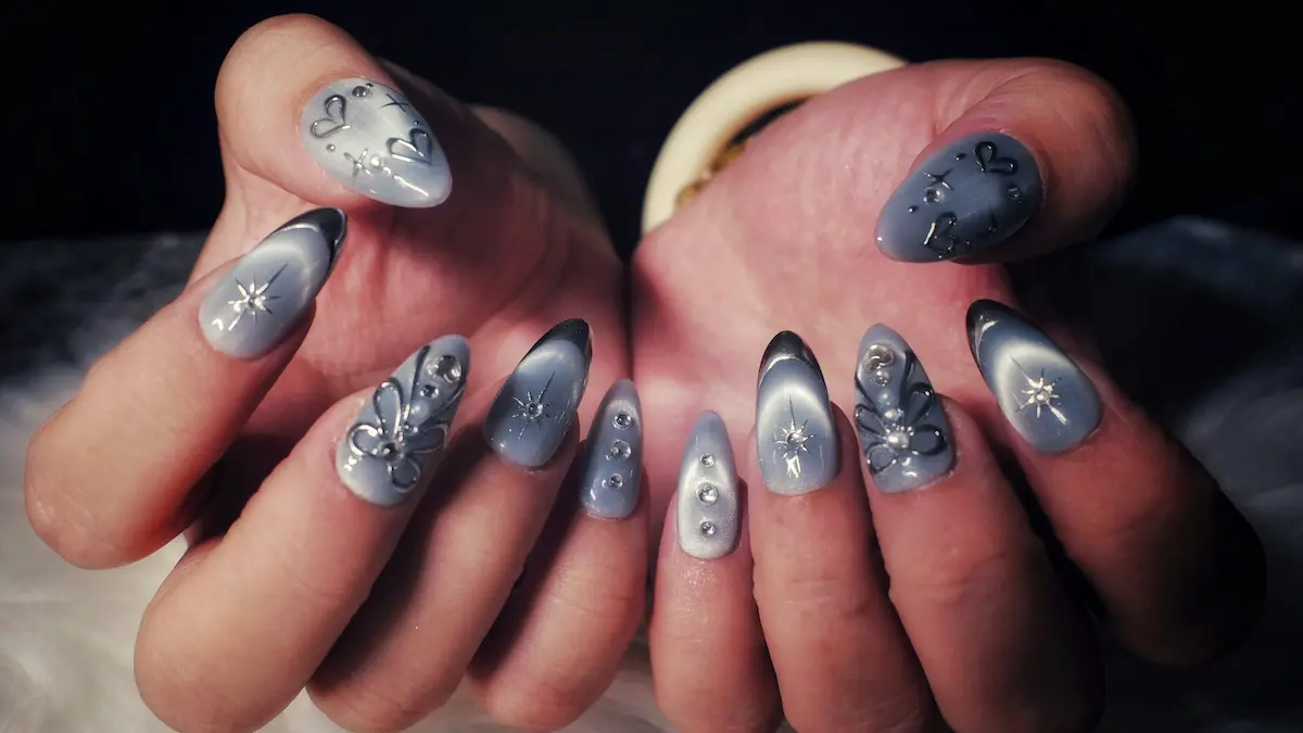 Grande plano das mãos de uma mulher com unhas de acrílico detalhadas, num design cinzento efeito olho de gato, com corações e estrelas prateados e aplicações de pedras brilhantes