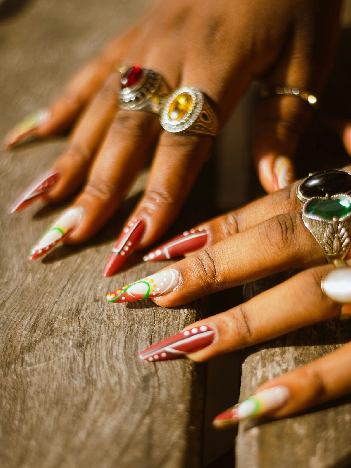 Grande plano das mãos de uma mulher com unhas de acrílico em formato stiletto, em vermelho e branco, decoradas com pintas e espirais verdes e amarelas
