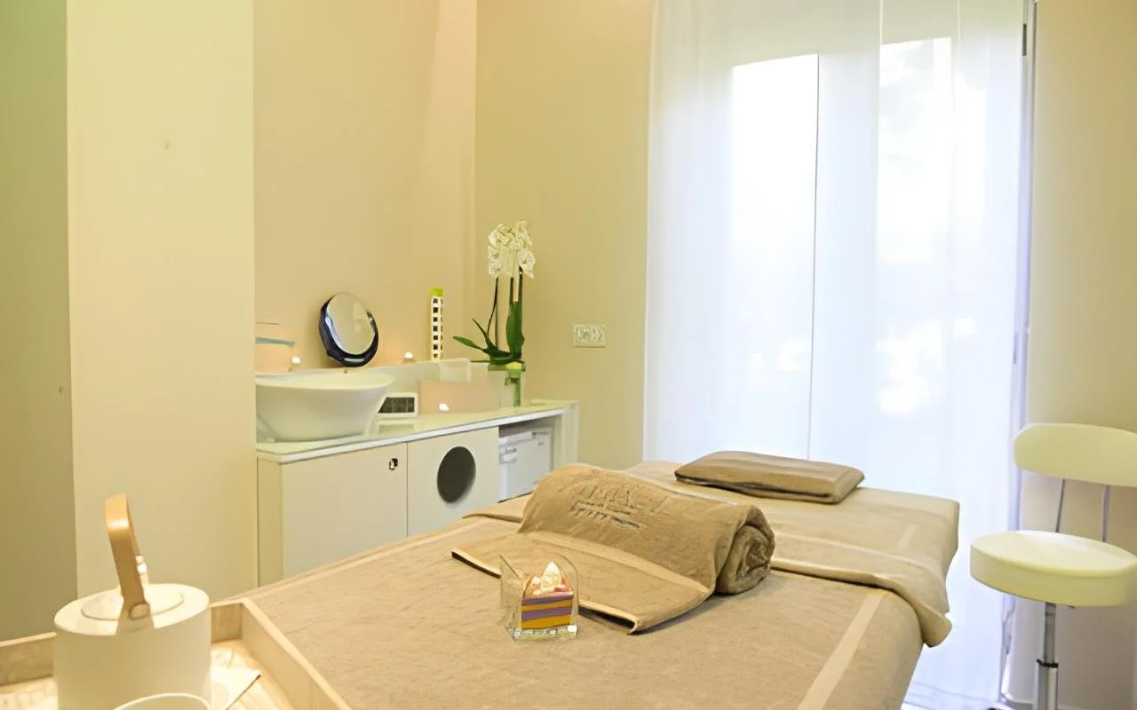 Uma sala de tratamento tranquila na Alice Beauty Clinique, com uma marquesa vestida de roupa de tom taupe suave, orquídeas brancas na bancada, velas junto ao lavatório e cortinas transparentes a filtrar a luz natural através de uma janela grande.
