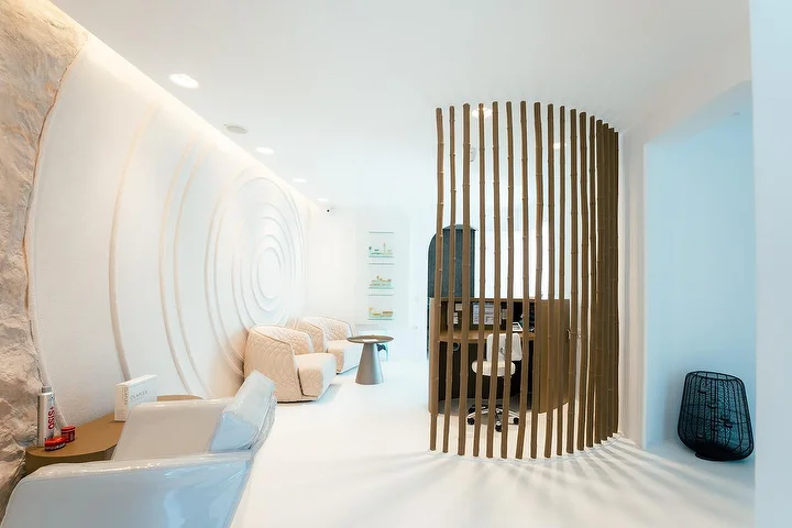 Um interior de spa tranquilo com ar de gruta, de paredes brancas curvas, uma delas esculpida em ondulações concêntricas, com iluminação indireta suave, poltronas acolchoadas cor de creme e um biombo curvo de ripas de madeira a delimitar um recanto de tratamento sossegado.
