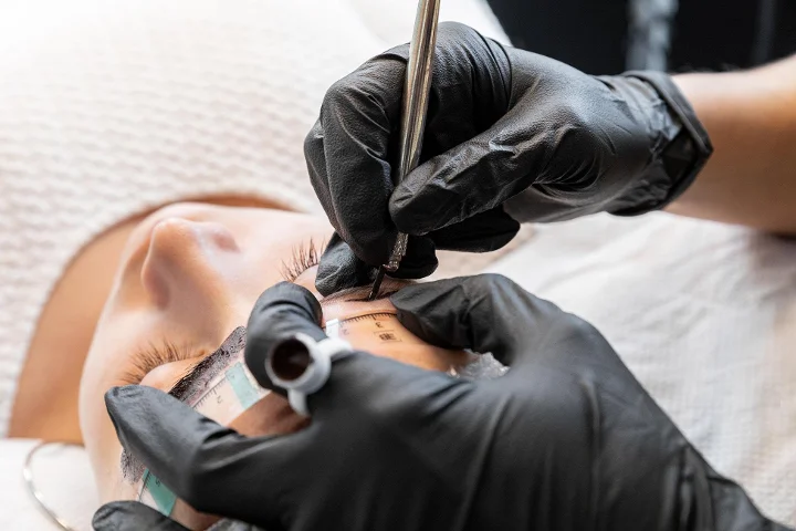 Grande plano de um tratamento de sobrancelhas em curso: uma técnica de luvas pretas mede e marca a sobrancelha de uma cliente reclinada com uma régua própria e um lápis, de olhos fechados.