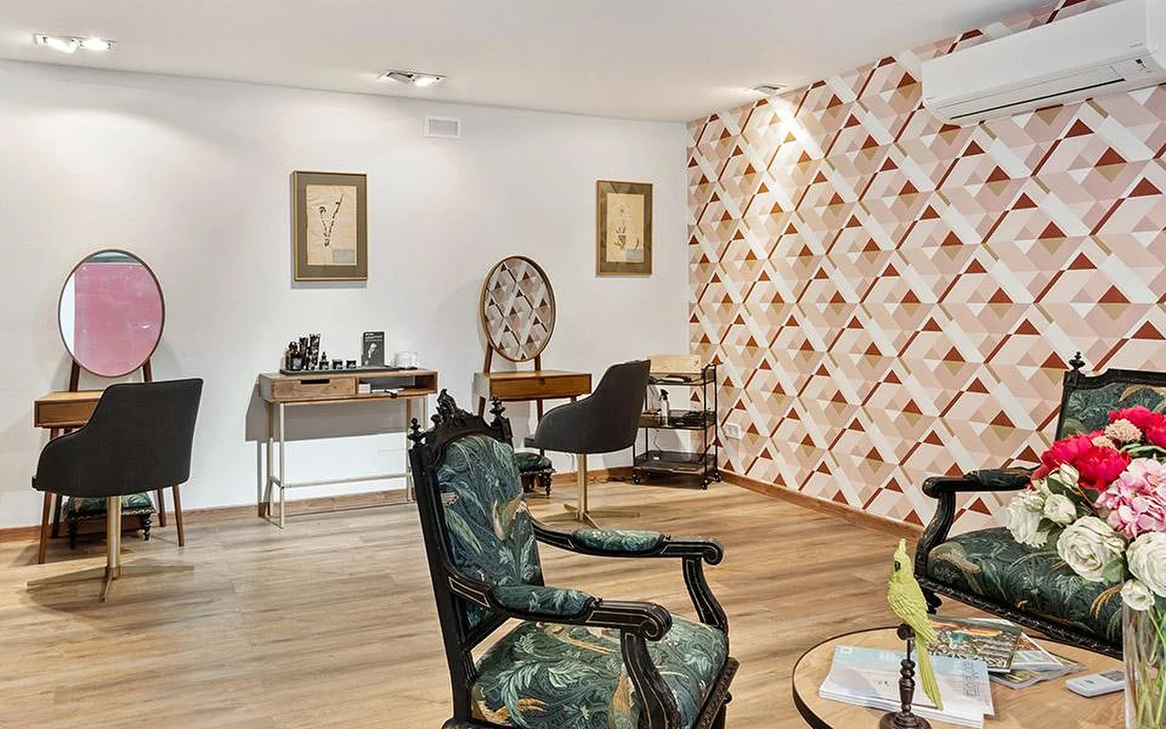 Uma sala de salão elegante com uma parede decorativa geométrica em rosa e terracota, um par de cadeiras entalhadas e ornamentadas forradas a tecido botânico verde, bancadas de styling em madeira com espelhos redondos e uma jarra de flores cor-de-rosa e vermelhas em primeiro plano.