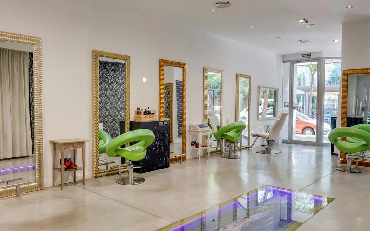 Um salão de cabeleireiro amplo e luminoso, com pavimento de betão polido, espelhos altos de moldura dourada ao longo de uma parede branca, cadeiras de styling moldadas em verde-lima e uma zona de chão de vidro iluminada a luz roxa, com a rua visível através das portas envidraçadas ao fundo.