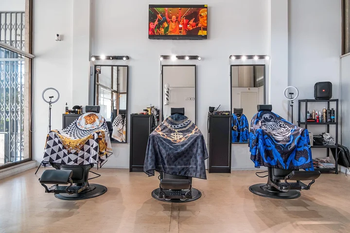 Uma barbearia ampla e luminosa de tetos altos com três cadeiras pretas cobertas por capas estampadas, cada uma frente a um espelho emoldurado por lâmpadas, com ring lights, um ecrã de parede e janelas altas.