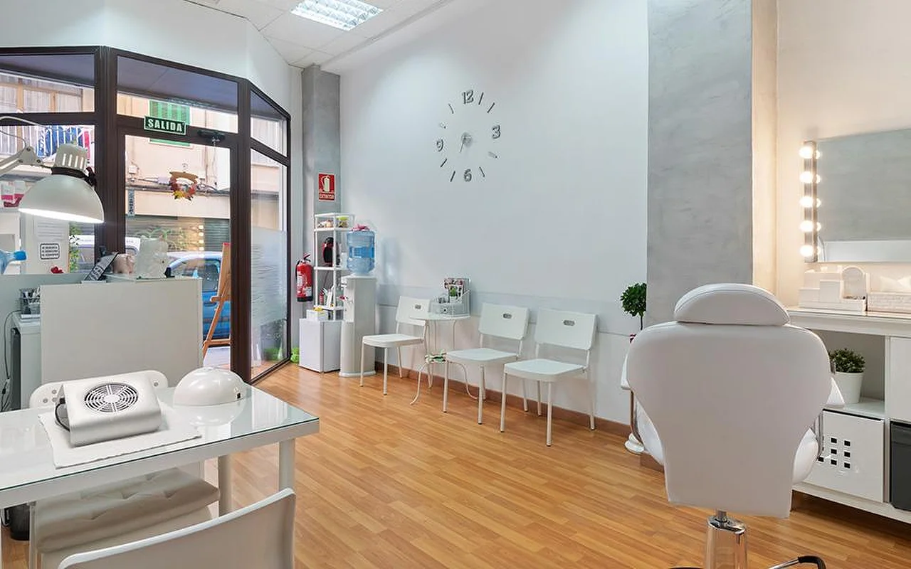 Um salão claro e branco, com pavimento de madeira clara, uma fila de cadeiras de espera brancas, um relógio de parede minimalista, um espelho de toucador iluminado, uma cadeira de tratamento reclinável branca e uma estação de manicure junto à janela.