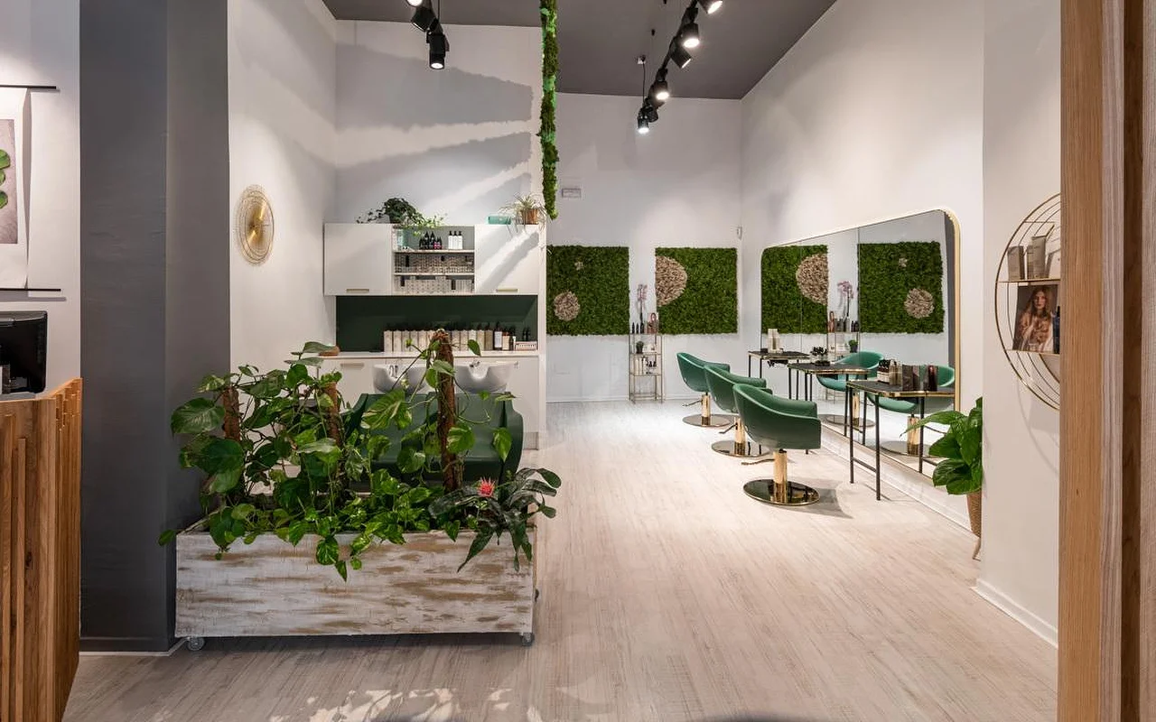 Um salão de cabeleireiro cheio de plantas, com pavimento de madeira clara, um vaso branco envelhecido a transbordar de verde, arte de parede em musgo verde, cadeiras de styling em veludo verde-esmeralda com detalhes dourados e uma zona de lavagem ao fundo.