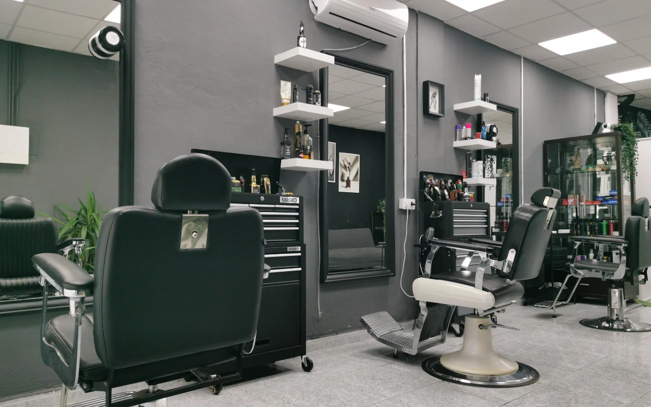 Uma barbearia tradicional com paredes cinzento-carvão, cadeiras de barbeiro clássicas em pele preta com bases e apoios de pés cromados, prateleiras suspensas brancas cheias de produtos de cuidado masculino e grandes espelhos sobre pavimento de azulejo.