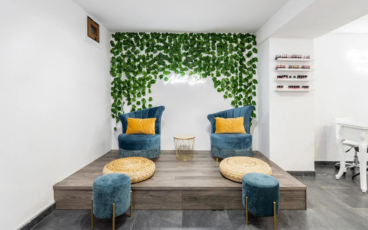 Um canto de nail bar com uma parede de folhagem verde em cascata e um pequeno letreiro de néon, duas poltronas de veludo azul-petróleo com almofadas mostarda sobre um estrado de madeira, pufes de descanso para os pés em fibra entrançada e prateleiras com vernizes coloridos à direita.