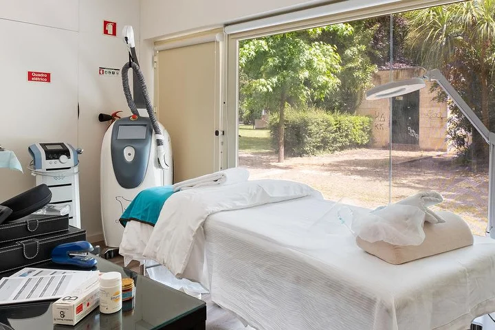 Uma sala de tratamentos com uma marquesa coberta de branco junto a uma janela do chão ao teto virada para um jardim verde, um aparelho de tratamentos de corpo num suporte, um candeeiro-lupa e uma mesa de vidro com produtos.