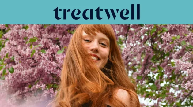 Retrato em primeiro plano de uma jovem ruiva sorridente com franja, com o cabelo a esvoaçar ao vento em frente a um fundo de flores de lilás cor-de-rosa. No topo, uma faixa azul-claro mostra o logótipo da "Treatwell" em azul-escuro.