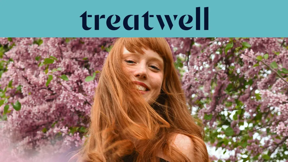 Retrato em primeiro plano de uma jovem ruiva sorridente com franja, com o cabelo a esvoaçar ao vento em frente a um fundo de flores de lilás cor-de-rosa. No topo, uma faixa azul-claro mostra o logótipo da 'Treatwell' em azul-escuro.