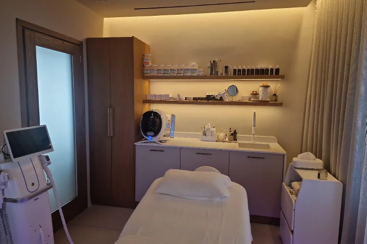 Uma sala de tratamento de luz quente, com uma marquesa facial branca e toalhas dobradas com cuidado, prateleiras de madeira escura cheias de produtos de cuidado da pele, um ecrã de análise da pele e aparelhos de tratamento ao lado, clínica mas acolhedora.