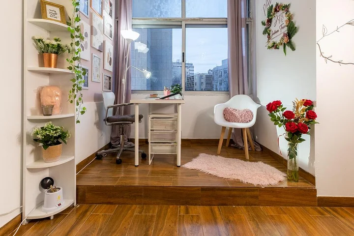 Um pequeno estúdio de estética sobre um estrado de madeira, com uma secretária branca e um candeeiro de pinça, uma cadeira de escritório e um cadeirão, prateleiras de canto com plantas, uma jarra de rosas vermelhas e uma janela sobre a cidade.