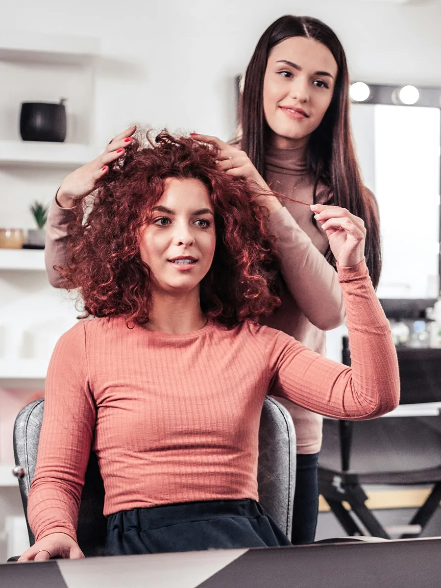 Uma cliente com cabelo cacheado pintado de vermelho sentada no cabeleireiro enquanto o profissional conversa com ela sobre o tratamento