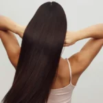Vista de trás de uma mulher com um top branco de alças, de braços levantados, a passar os dedos pelo seu cabelo castanho, comprido e brilhante