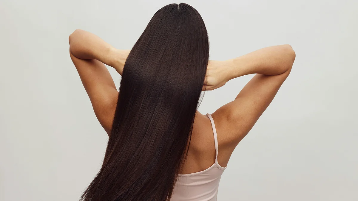 Vista de trás de uma mulher com um top branco de alças, de braços levantados, a passar os dedos pelo seu cabelo castanho, comprido e brilhante