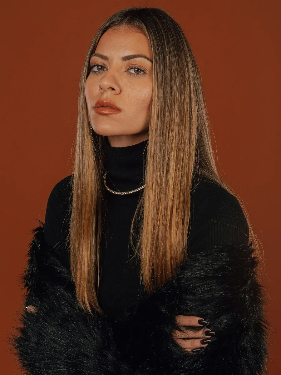 Retrato de estúdio de uma mulher com cabelo castanho-médio, comprido e brilhante, com um top preto, uma estola de pelo preta e argolas e colar brilhantes