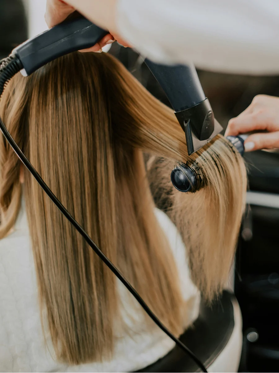 Vista de trás de uma mulher num cabeleireiro enquanto o profissional lhe seca o cabelo liso durante uma sessão de tratamento de queratina