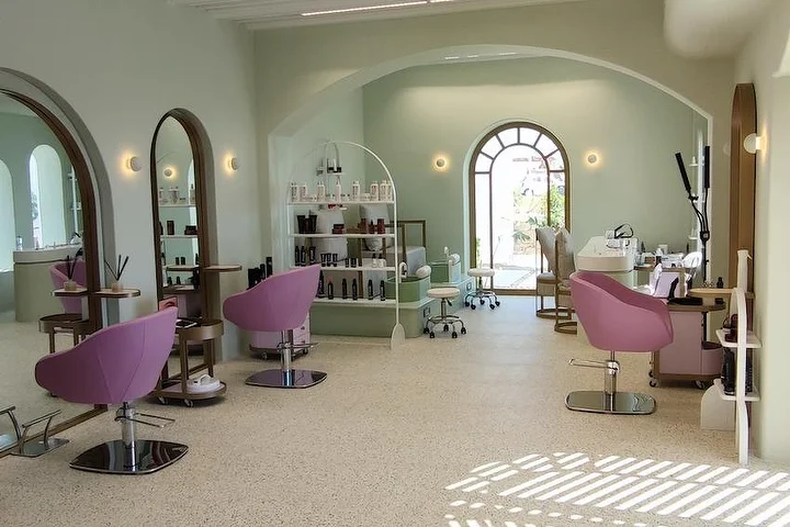Um salão luminoso em verde-salva com pavimento em terrazzo, espelhos em arco com moldura de madeira e uma janela em arco para o jardim, cadeiras de styling em rosa-velho e prateleiras abertas cheias de produtos, com a luz do sol a cair sobre o chão.