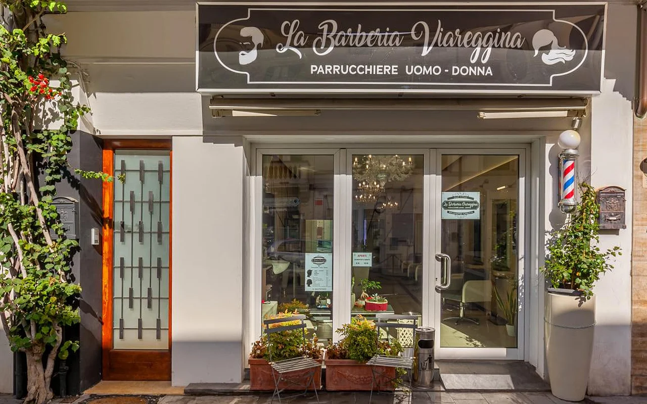 A montra da La Barberia Viareggina num dia de sol, com um clássico poste de barbeiro vermelho, branco e azul à direita das portas de vidro. Plantas em vasos e trepadeiras enquadram a entrada, e vê-se um candeeiro de teto lá dentro através das janelas.