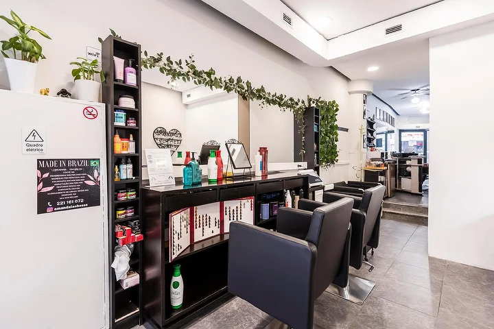 Um cabeleireiro estreito e comprido com cadeiras pretas ao longo de uma parede de espelhos, uma prateleira escura cheia de produtos de cabelo, uma grinalda de folhas a descer sobre o espelho e uma placa emoldurada de Made In Brazil numa coluna branca.