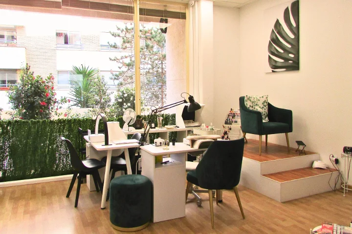 Um espaço de manicure luminoso com duas mesas de unhas brancas e respetivos candeeiros, cadeiras de veludo verde e um banco a condizer, uma tela com folha de monstera na parede e janelas grandes para um terraço com plantas.