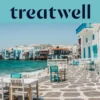 A marginal da Pequena Veneza em Mykonos: casas caiadas de branco com varandas de madeira coloridas à beira da água, o mar Egeu turquesa ao fundo e, em primeiro plano, mesas de café brancas e verde-azuladas dispostas num terraço de pedra.