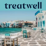A marginal da Pequena Veneza em Mykonos: casas caiadas de branco com varandas de madeira coloridas à beira da água, o mar Egeu turquesa ao fundo e, em primeiro plano, mesas de café brancas e verde-azuladas dispostas num terraço de pedra.