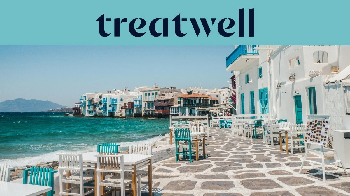 A marginal da Pequena Veneza em Mykonos: casas caiadas de branco com varandas de madeira coloridas à beira da água, o mar Egeu turquesa ao fundo e, em primeiro plano, mesas de café brancas e verde-azuladas dispostas num terraço de pedra.