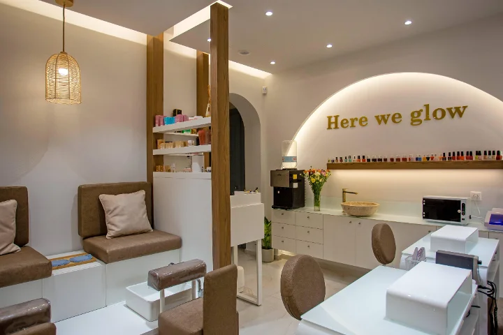 Um salão de unhas quente e de luz suave, com um candeeiro de teto em rotim entrançado, bancos de pedicure castanhos almofadados, mesas de manicure brancas, uma prateleira de vernizes coloridos e um letreiro dourado «Here we glow» sobre um arco retroiluminado.