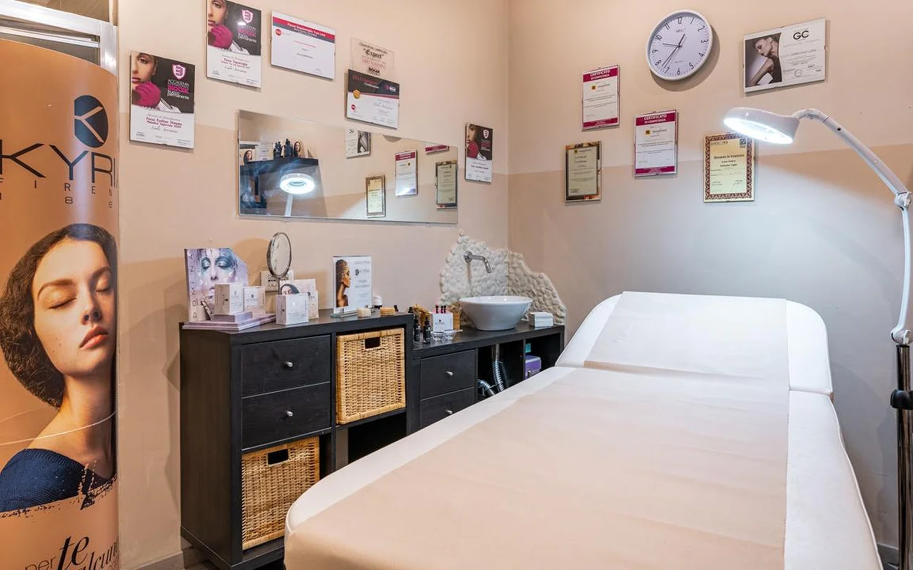 Uma sala de tratamento na Oltre la bellezza com uma marquesa branca, um candeeiro com lupa ajustável e paredes forradas de certificados e diplomas profissionais. Móveis de arrumação em vime escuro ficam por baixo de um espelho, com produtos de cuidado da pele dispostos na bancada.