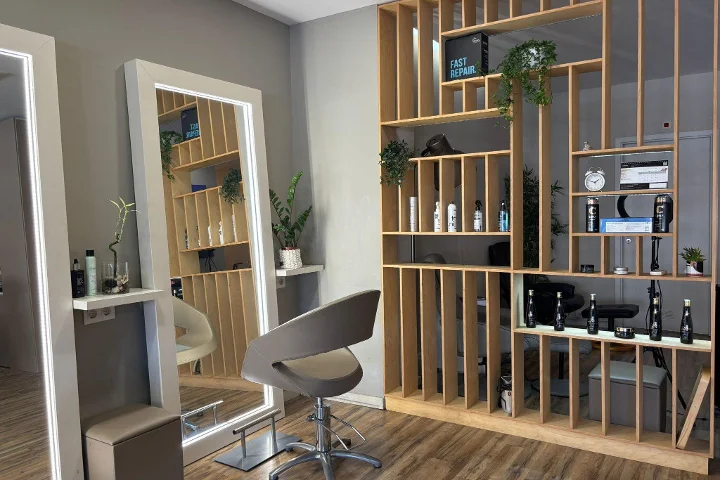 Uma estação de cabeleireiro com uma cadeira cinzenta em frente a um espelho emoldurado e iluminado por LED, uma estante-divisória de ripas de madeira com produtos de styling, um relógio e plantas, sobre um chão com aspeto de madeira.