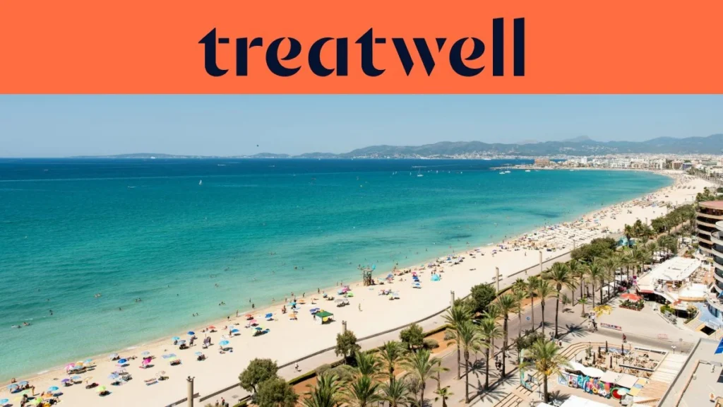 Vista aérea da Playa de Palma, em Maiorca, com uma longa curva de areia dourada, filas de chapéus de sol, água turquesa e calma e uma avenida marginal ladeada de palmeiras junto à cidade, com colinas ao fundo sob um céu azul e limpo.